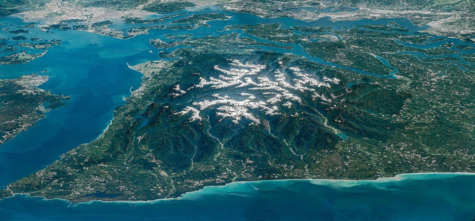 The Olympic Peninsula, viewed at an angle from above, features snow-capped mountains surrounded by deep, forested river valleys. The islands of Puget Sound and developed areas such as Seattle and Tacoma appear at the top of the photo.