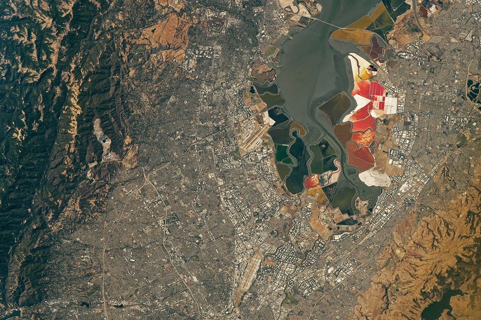 An aerial view of San Francisco Bay Area Stadium, center left, is surrounded by urban infrastructure. Towards the top you can see colorful pools of the southern San Francisco Bay.
