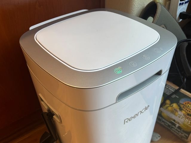 Side view of the Reencle Prime electric home composter, a white rectangular device with a small black scoop attached to the side