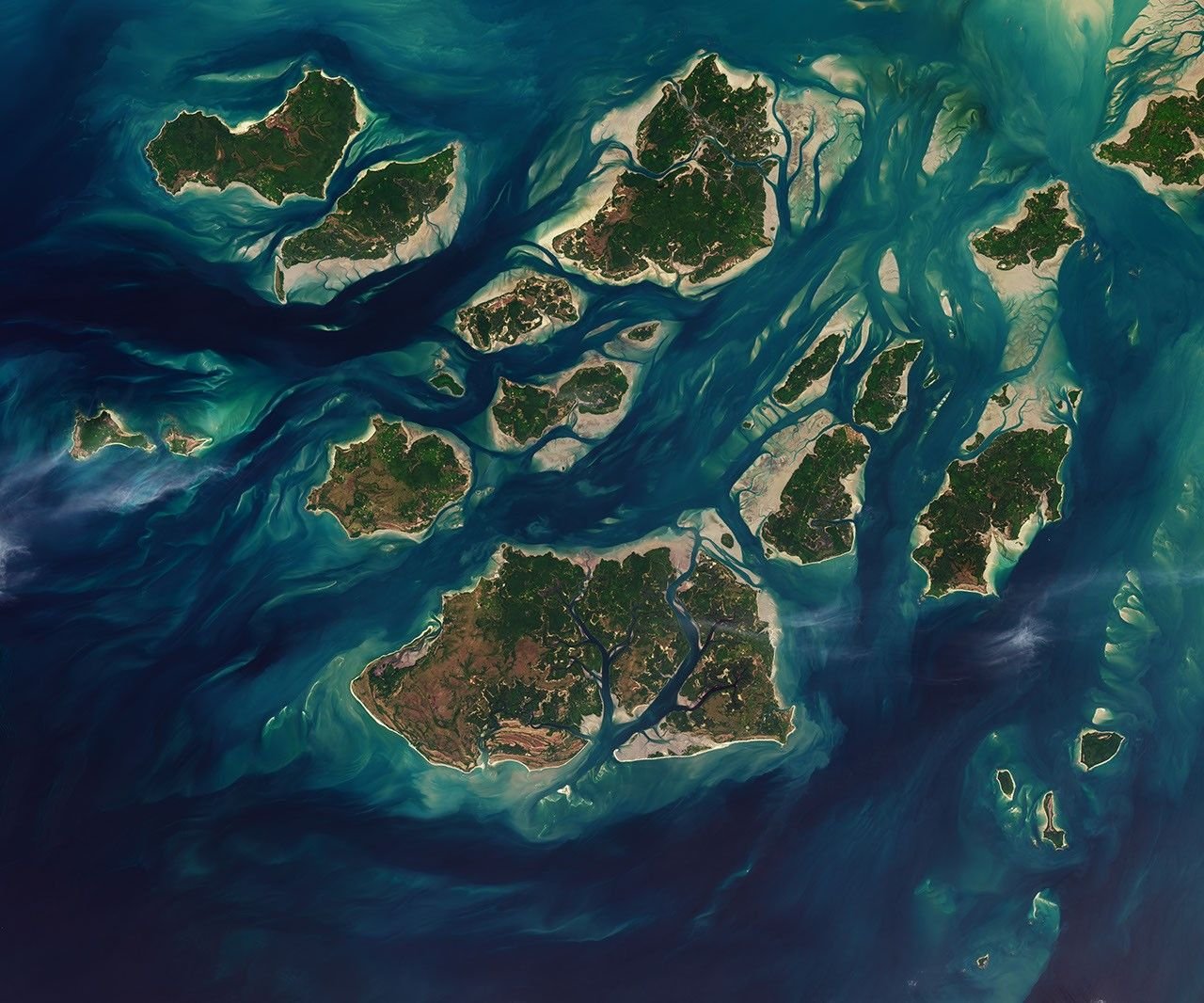 A satellite image shows a group of green islands surrounded by beige sand plains and networks of canals filled with dark blue water.