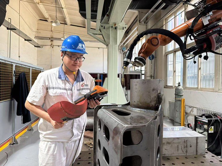 Wu Xiangyang wears a blue helmet and holds a welding protective helmet. He is standing in front of a robotic welding arm in the CRRC factory.