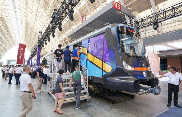 People climb a display of a new subway train up stairs in a large, bright hangar. In the background, a large group of people gather under a red flag with the letters CRRC.