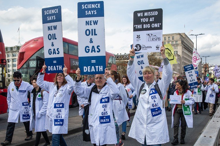 Scientists in lab coats march across Waterloo Bridge in London, holding signs to protest the fossil fuel industry.