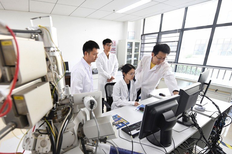 Three young Chinese scientists in lab coats, looking at a computer monitor, one of them is sitting and there is a fourth older scientist offering advice.
