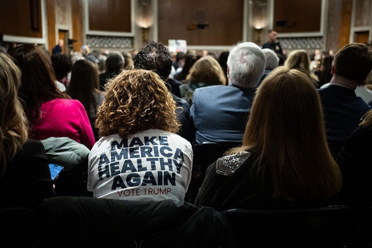 An audience sits in a large Senate hearing room, and an attendee wears a white T-shirt emblazoned with the slogan “MAKE AMERICA HEALTHY AGAIN – VOTE TRUMP” as a speaker addresses the crowd at the front.