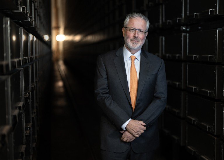 Portrait of Neil Shubin standing in a dimly lit, side-lit, wood-paneled hallway.