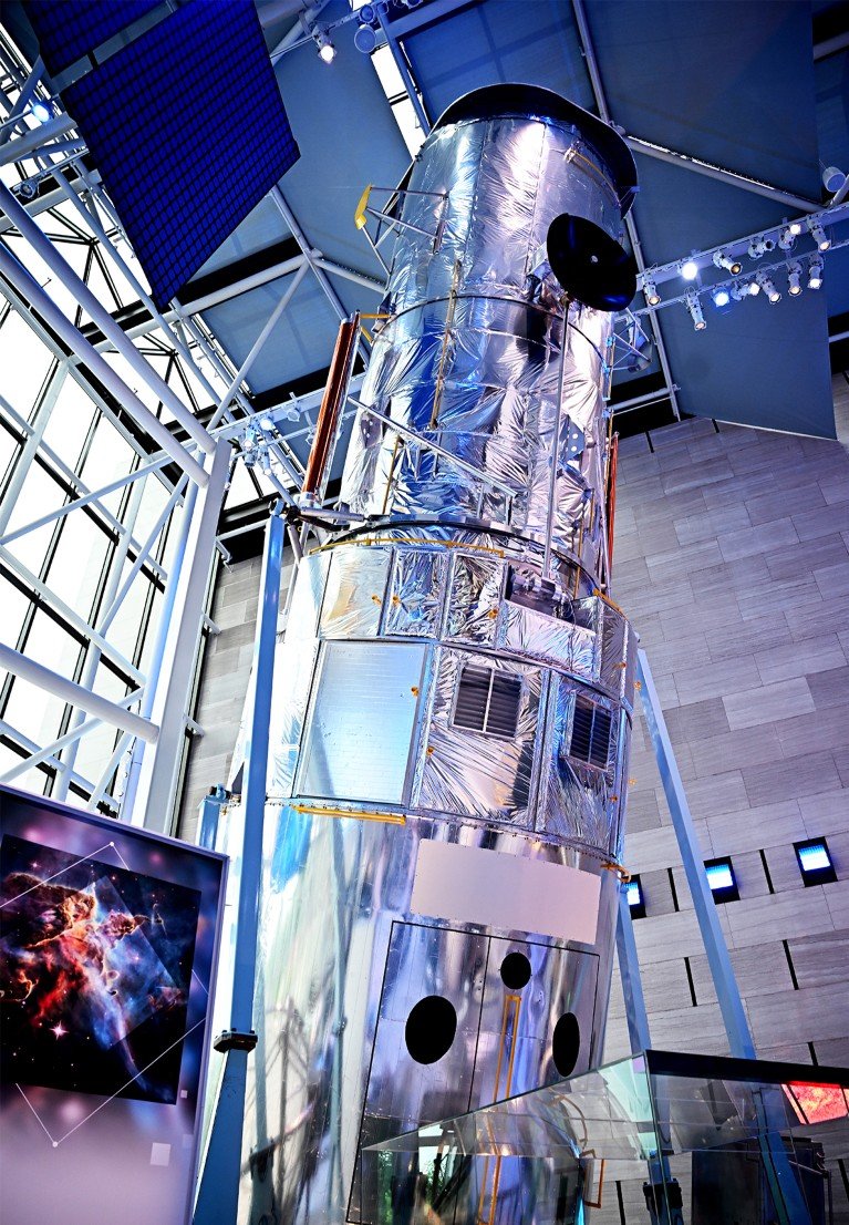 A large cylindrical space telescope model with metallic reflective surfaces and various structural components.