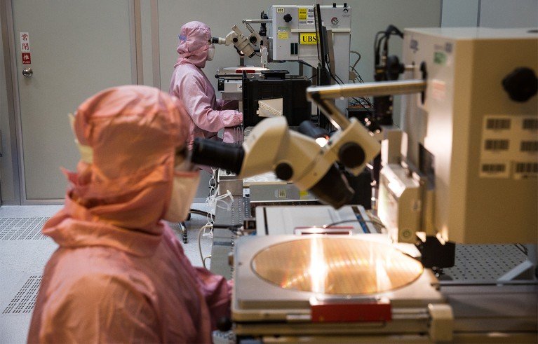 Two scientists dressed in light pink clean room suits operate high-powered microscopes to inspect large circular silicon wafers used in the manufacturing of semiconductor microchips.
