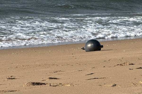 A gray sphere shines in the sun on the sand of a beach, with the sea in the background.