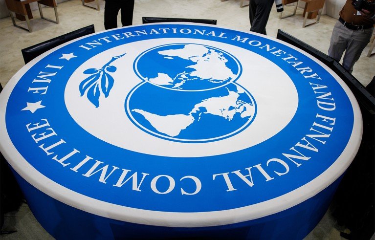 A large circular table with the International Monetary Fund (IMF) logo highlighted in blue and white in a meeting place. The logo features a world map and laurel branches.