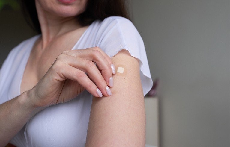 A woman wearing a white blouse and applying a small hormone replacement adhesive patch to her upper arm.