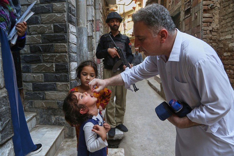 On the steps of a gate, a health worker administers a polio vaccine to a child while an armed police officer stands guard.