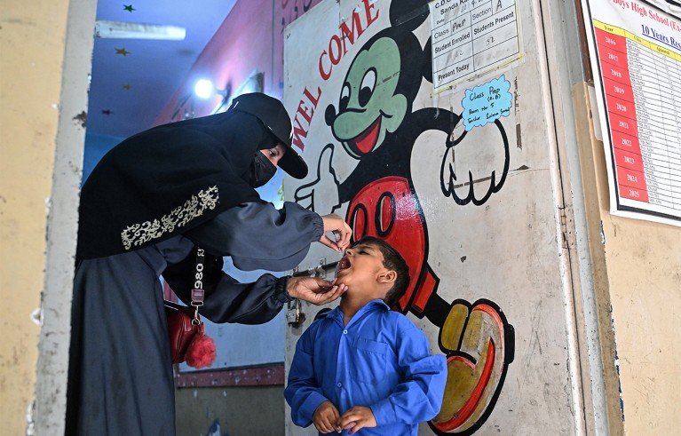 A woman in a black niqab administering oral medicine to a child in a blue school uniform inside a classroom. A large, colorful Mickey Mouse mural with a "WELCOME" The sign is painted on the wall behind them, along with educational posters and graphics.