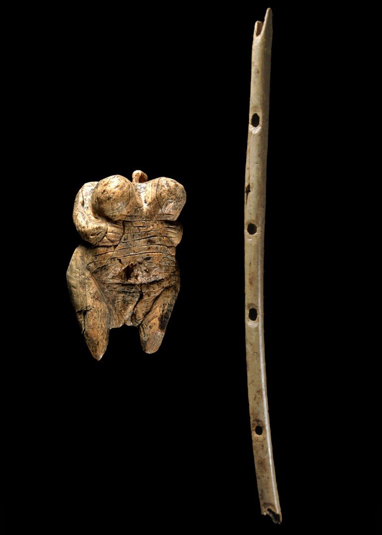 A composite image of a small carved female figure and a thin bone flute with finger holes on the right, on a black background