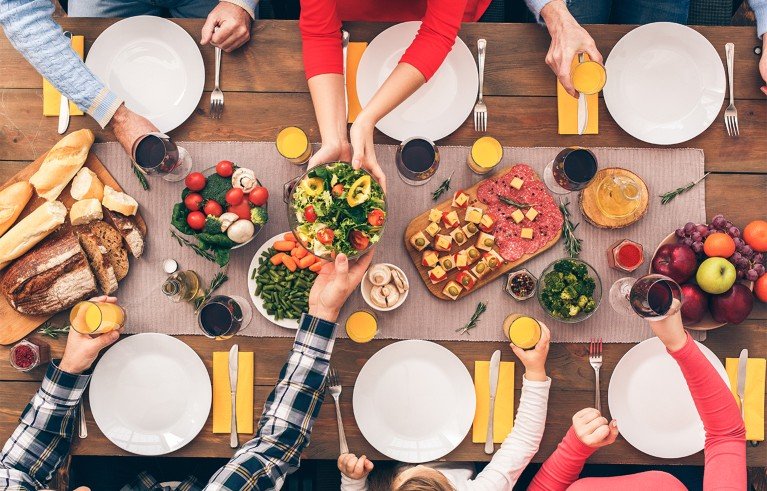 A top-down view of a festive dining table set for six people with empty white plates, yellow napkins and various plates including salad, bread, cold cuts and fruit. Hands of six people reach out to serve or hold glasses of orange juice and red wine.