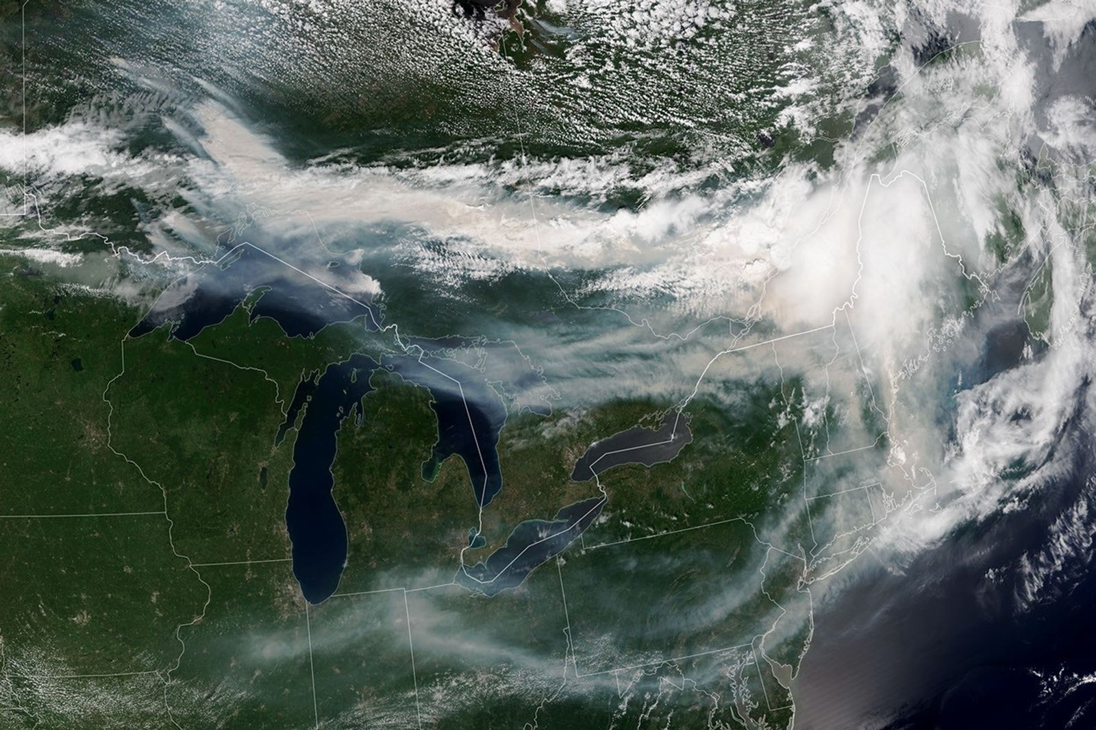 A satellite image shows brown smoke from wildfires in Ontario, Canada, moving east across parts of Canada and the United States. Areas of white clouds mix with the smoke.
