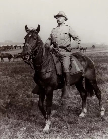 Teddy Roosevelt on horseback. He wears a wide-brimmed hat and glasses