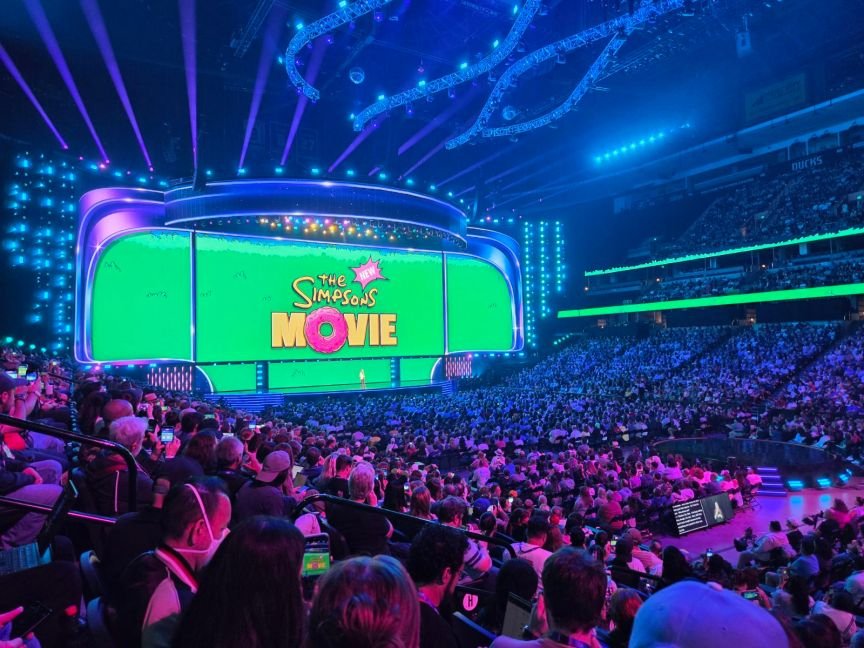 A large screen with the words "The Simpsons New Movie" in the animated series' iconic font in front of a large crowd of people.