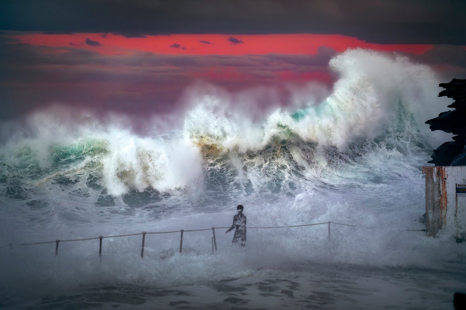 A teenager stands as waves crash over fence