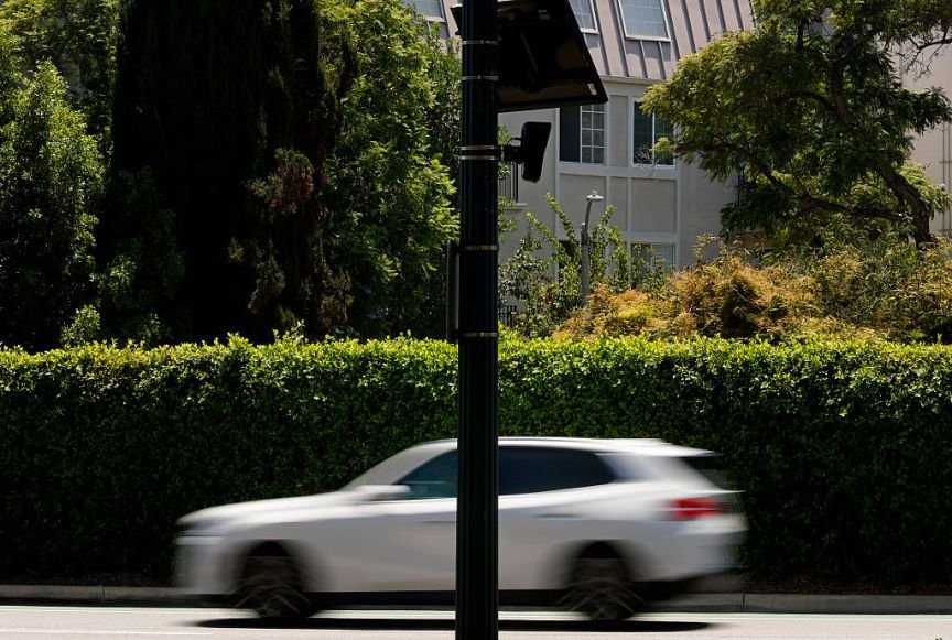 A white car passing by a shrub and Flock camera.