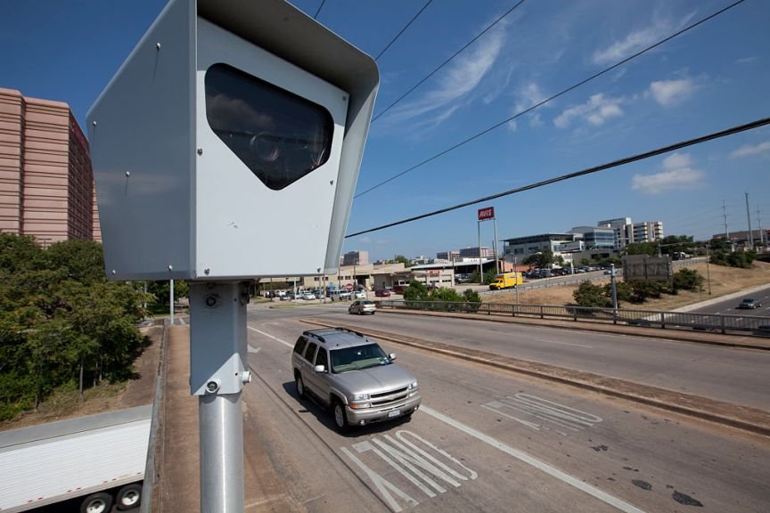 A white, boxy red light camera next to a highway.