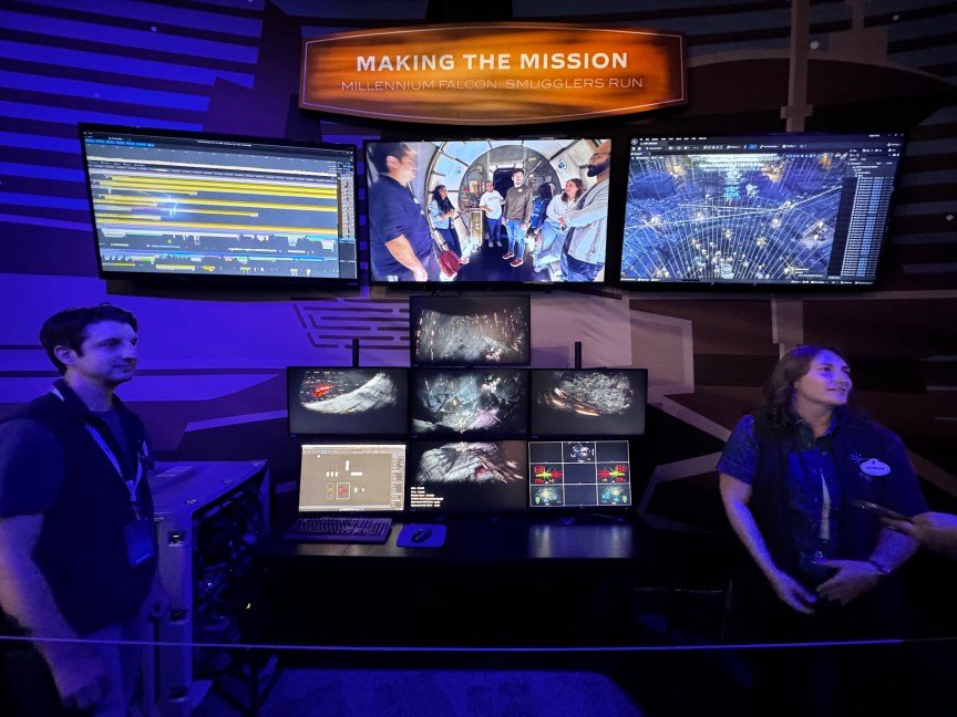two people stand next to a control console with several screens