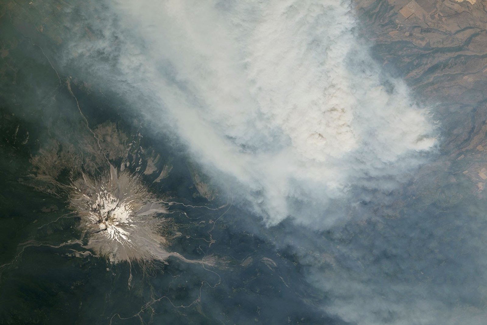 An ice-capped volcano stands in the bottom-left corner, with a wide plume of brown-gray smoke filling most of the rest of the frame.
