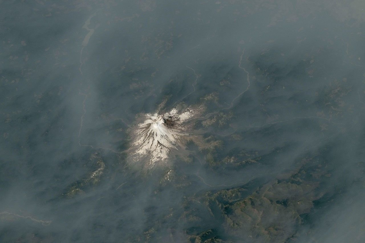 An ice-capped volcano stands at the center of the image, its surrounding terrain blanketed in brown-gray smoke.
