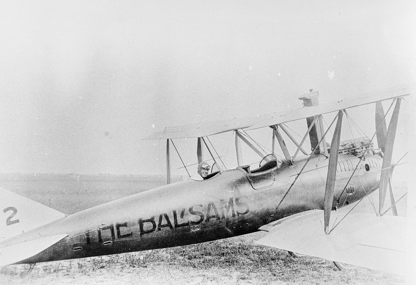 a black and white photo of a pilot sitting in the back seat of a biplane in 1920. the plane has "the balsams" written on it
