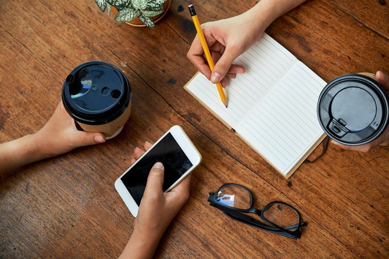 Close-up view of a table where two people are meeting. It shows their hands — one person is holding a smart phone and a takeaway cup of coffe and the other makes notes in a notebook with a pencil while also holding a takeaway coffee.