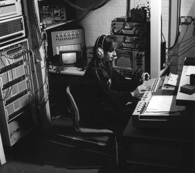 A black and white archive photo of Susan James at work in the RAE acoustics laboratory, wearing a headset and typing on a word processor, circa 1980.
