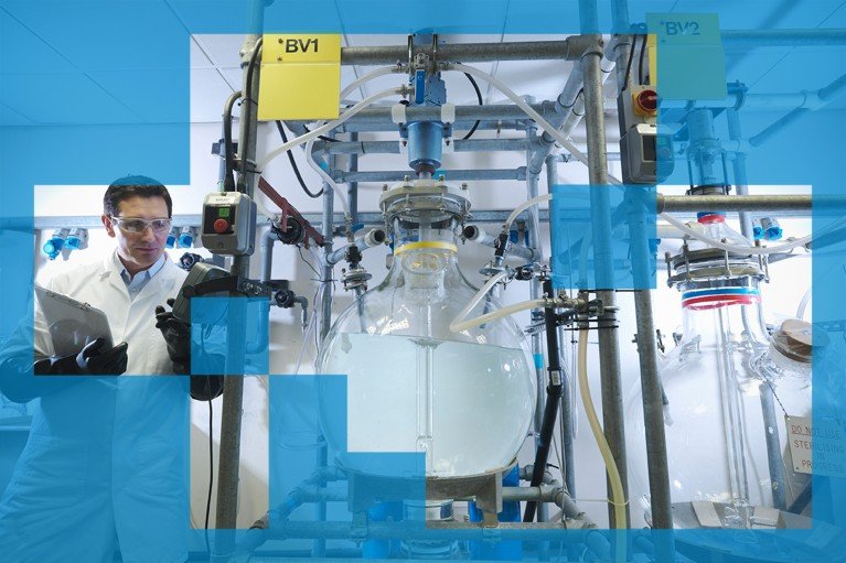 A laboratory professional supervising a large glass reactor vessel connected to an extensive network of pipes, valves, sensors, and control equipment in a research environment.