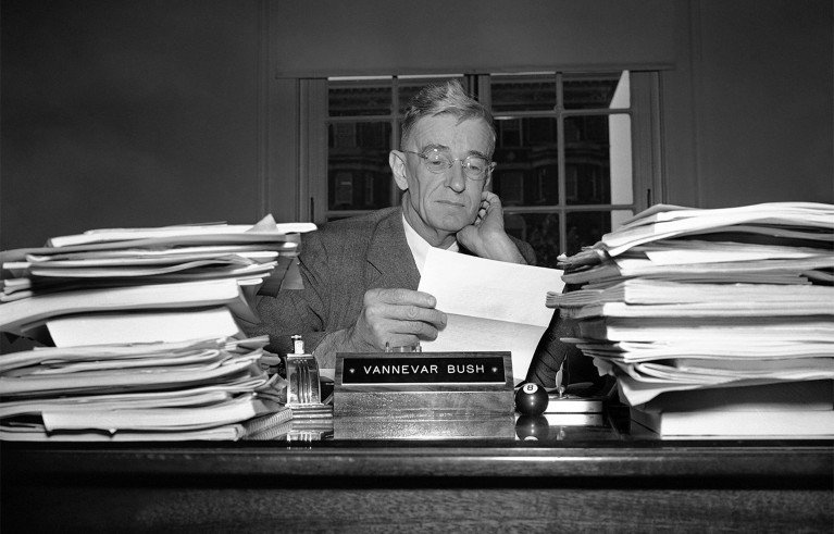 Black and white photograph of Vannevar Bush sitting at a desk cluttered with large stacks of papers on both sides. He is reading a document with a focused expression.