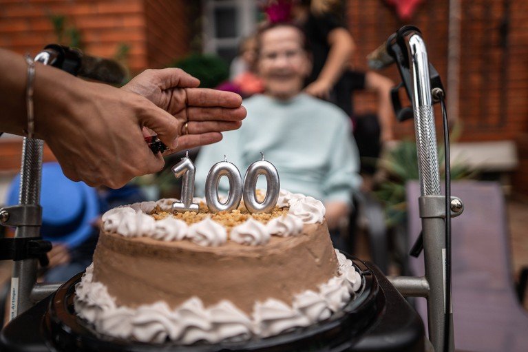 A chocolate and cream birthday cake with a '100' candle ready to be lit by a person out of shot. In the diffuse background an elderly woman is seated looking on.