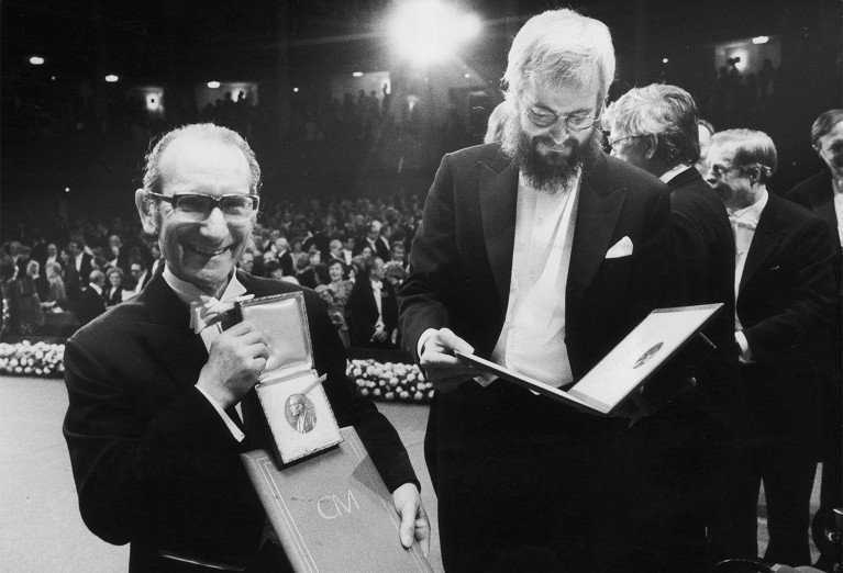 Black and white photograph of two men in formal attire at a ceremony in front of an audience. One man is holding a medal and certificate while smiling at the camera.