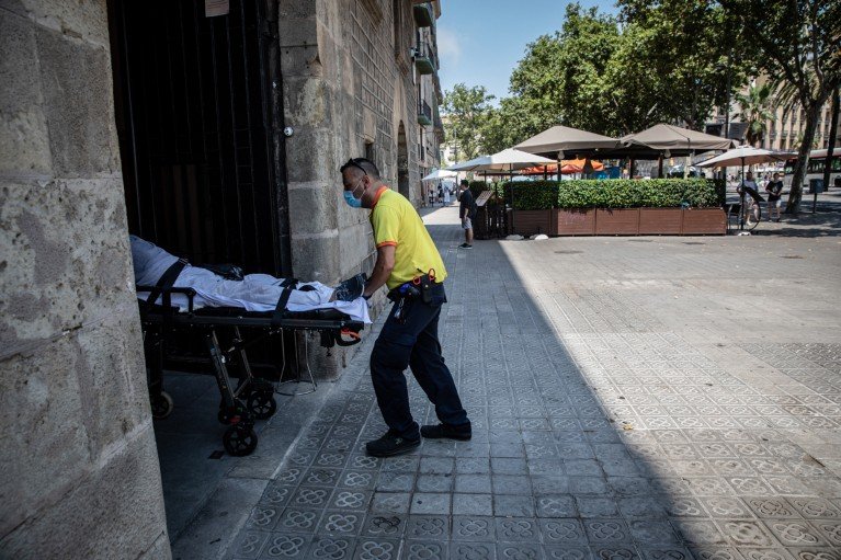A person wearing a high-visibility yellow shirt and a face mask pushes a patient under a white blanket on a wheeled stretcher through a large stone doorway on a city street.