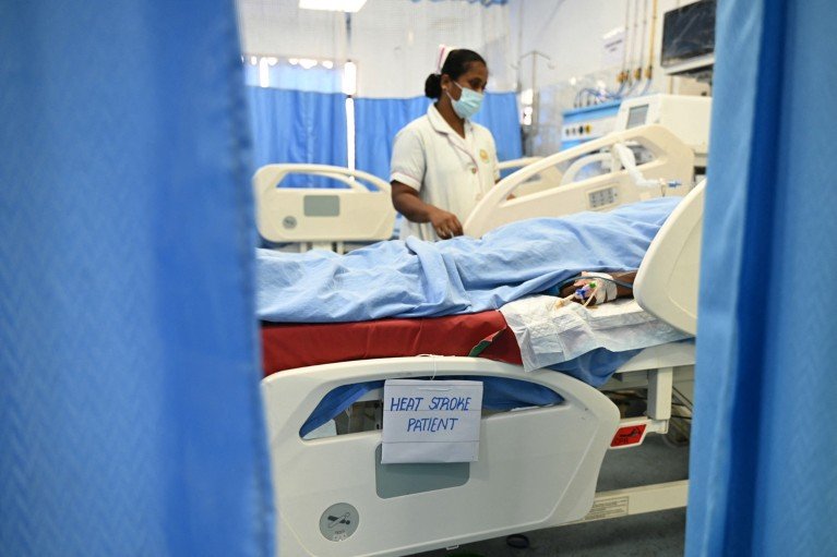 A patient lies in a hospital bed under a blue sheet, with a healthcare worker nearby and a sign attached to the bed reading “heat stroke patient”