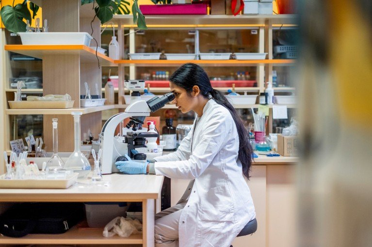 A person in a white lab coat and blue gloves sits at a laboratory bench looking down a microscope