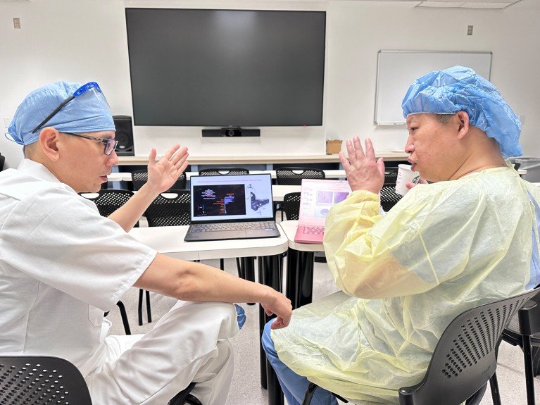 Two men wearing surgical caps and protective clothing sit in a training room, discussing the information displayed on laptops in front of them.