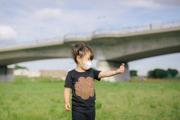 A masked girl plays in a grassy park while holding a flower, with a cement footbridge in the background.