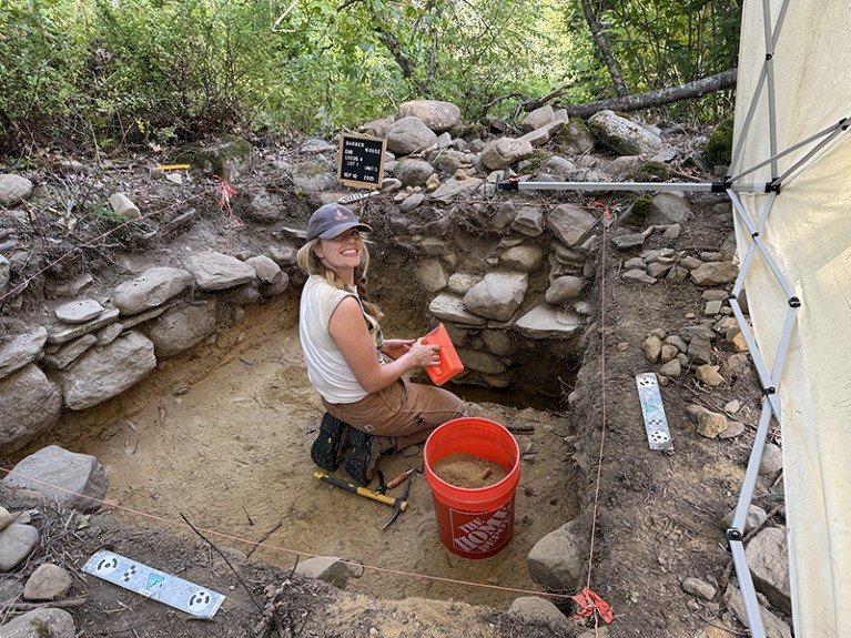 Jasper Clayton kneeling in a square pit lined with stones during the excavation of a Colonial cellar hole in Pemaquid, Maine. She holds a red plastic scoop and kneels next to a large bucket full of soil and other archaeological digging tools.
