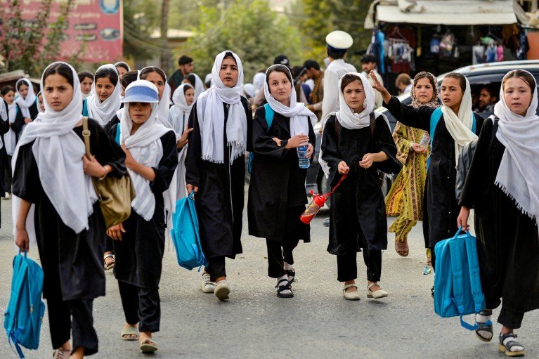 Girls in dark uniforms and white headscarves walk along a busy street carrying blue backpacks.