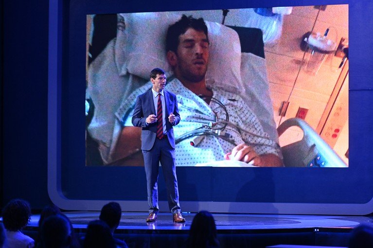 David Fajgenbaum speaking from a conference hall stage. Behind him the presentation screen shows a photo of himself laying in a hospital bed.