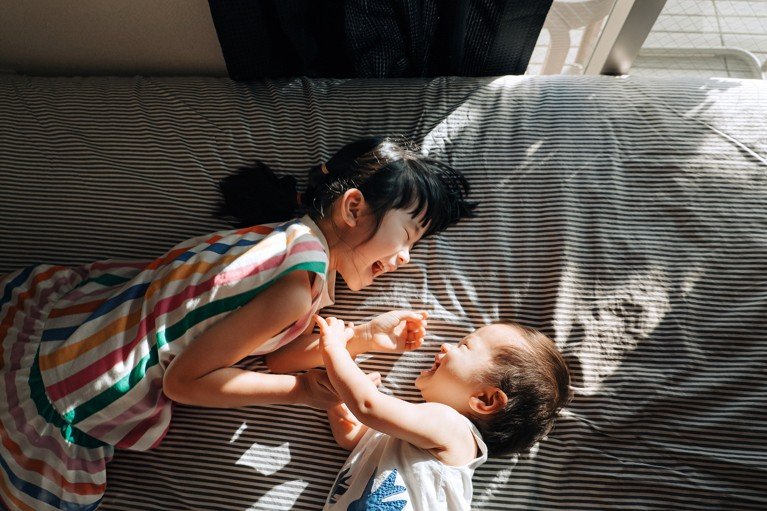 Asian girl laying on a bed with her toddler sibling laughing and tickling each other.