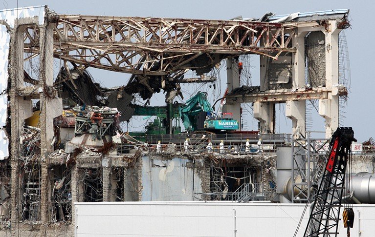 several people wearing hard hats and white protective clothing are visible walking on a damaged reactor building.