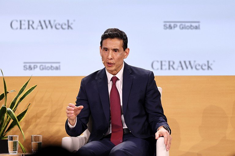 Ho Nieh is seen sitting in a chair, wearing a suit, shirt and tie. He is talking into a microphone attached to his head.