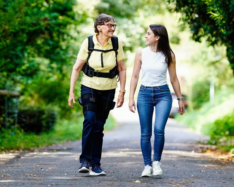 A senior citizen wearing exoskelton shorts walks beside a younger woman through a park.