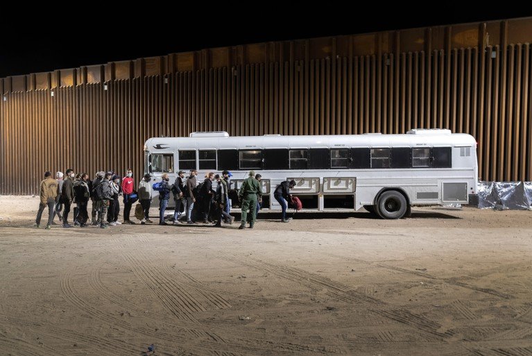 A group of people queue next to a white bus parked on a dirt road to load their bags and belongings into the storage compartments at night