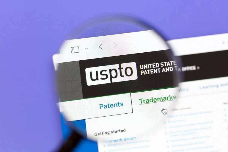 A magnifying glass in front of a laptop screen, focussing on the The United States Patent and Trademark Office USPTO logo from the webpage.
