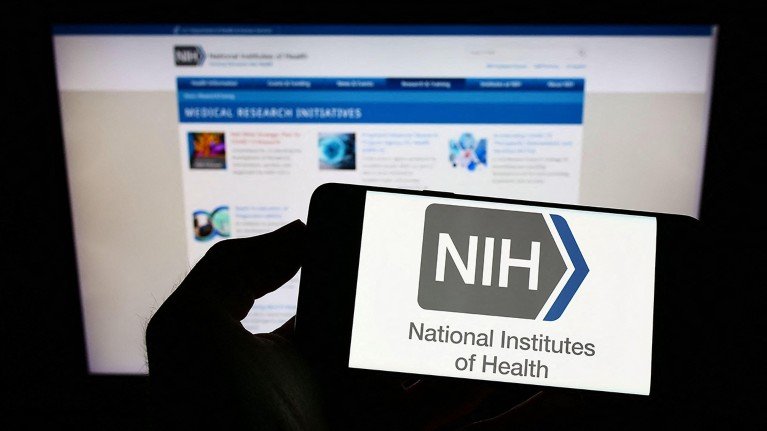 Hand holding a smartphone displaying the NIH logo in front of the NIH website, with focus on the phone screen.